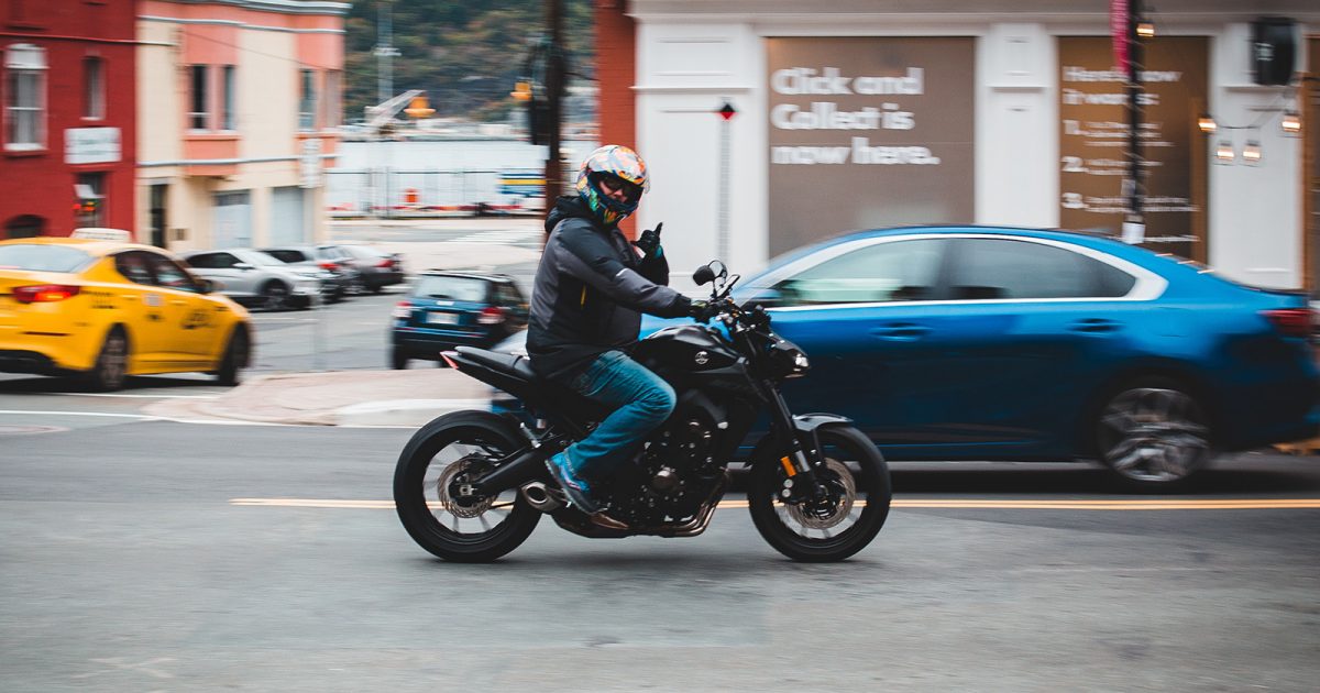 Hombre con casco conduce moto en la calle, detrás hay autos pasando.