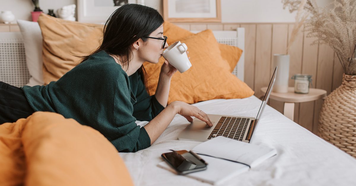Mujer tomando café, sobre la cama, con el ordenador, contratando un seguro online.