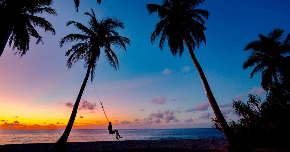foto de un atardecer en la playa, al centro se ve una persona descansando en una hamaca rodeado de palmeras